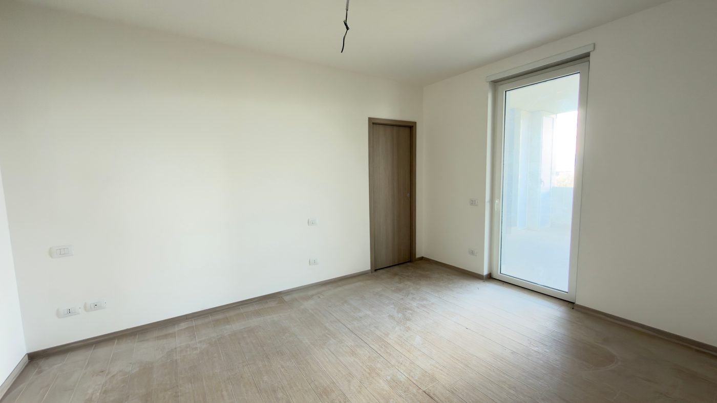 Prima: la foto originale, scattata col telefono, di una camera vuota, con pavimento in legno chiaro, pareti bianche, una porta in legno e una portafinestra.