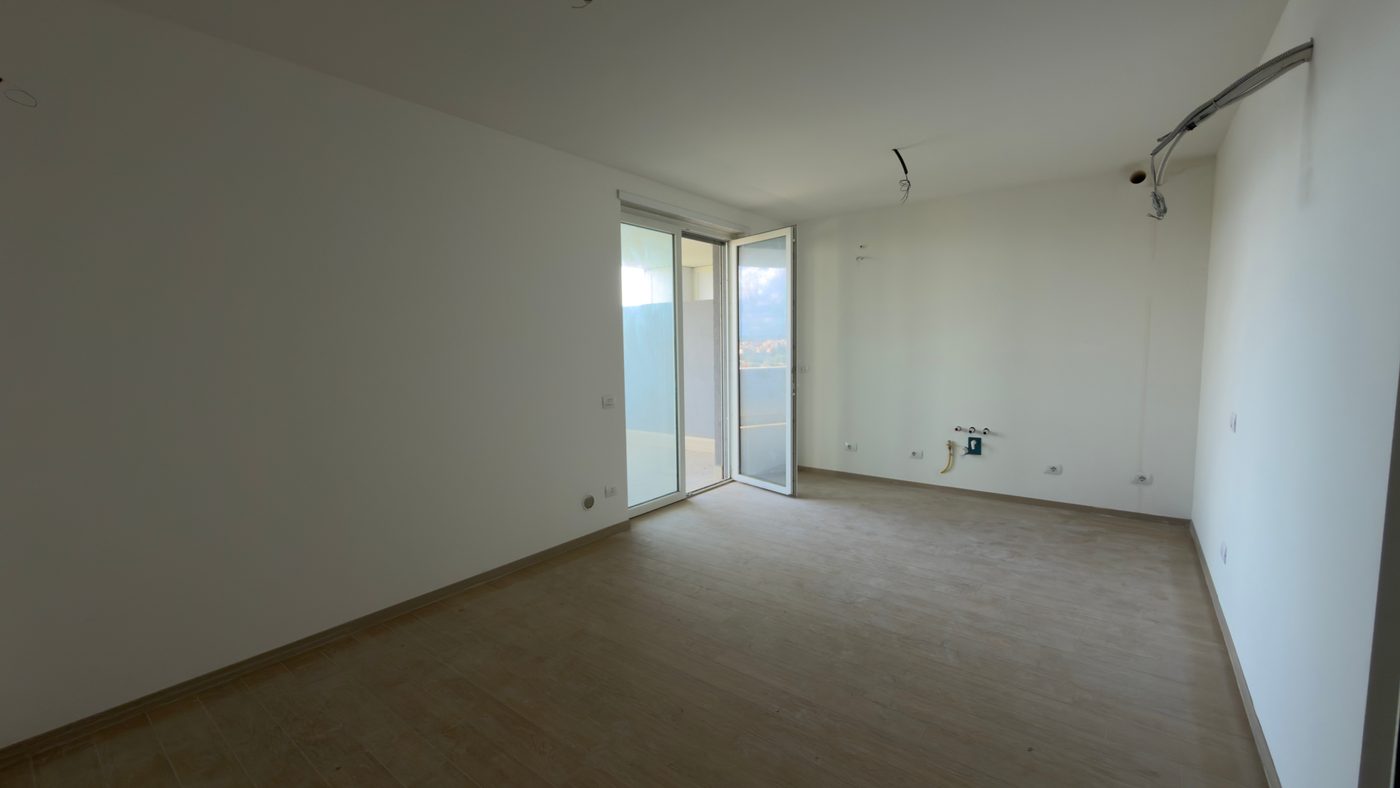 Prima: la foto originale, scattata col telefono, di una cucina con soggiorno vuota in cantiere: sulla parete di fondo solo gli attacchi di acqua e gas, a sinistra una portafinestra aperta sul balcone.